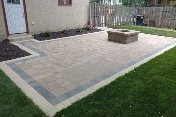 New tan paver patio with a gray border and square stone fire pit beside a fresh green lawn