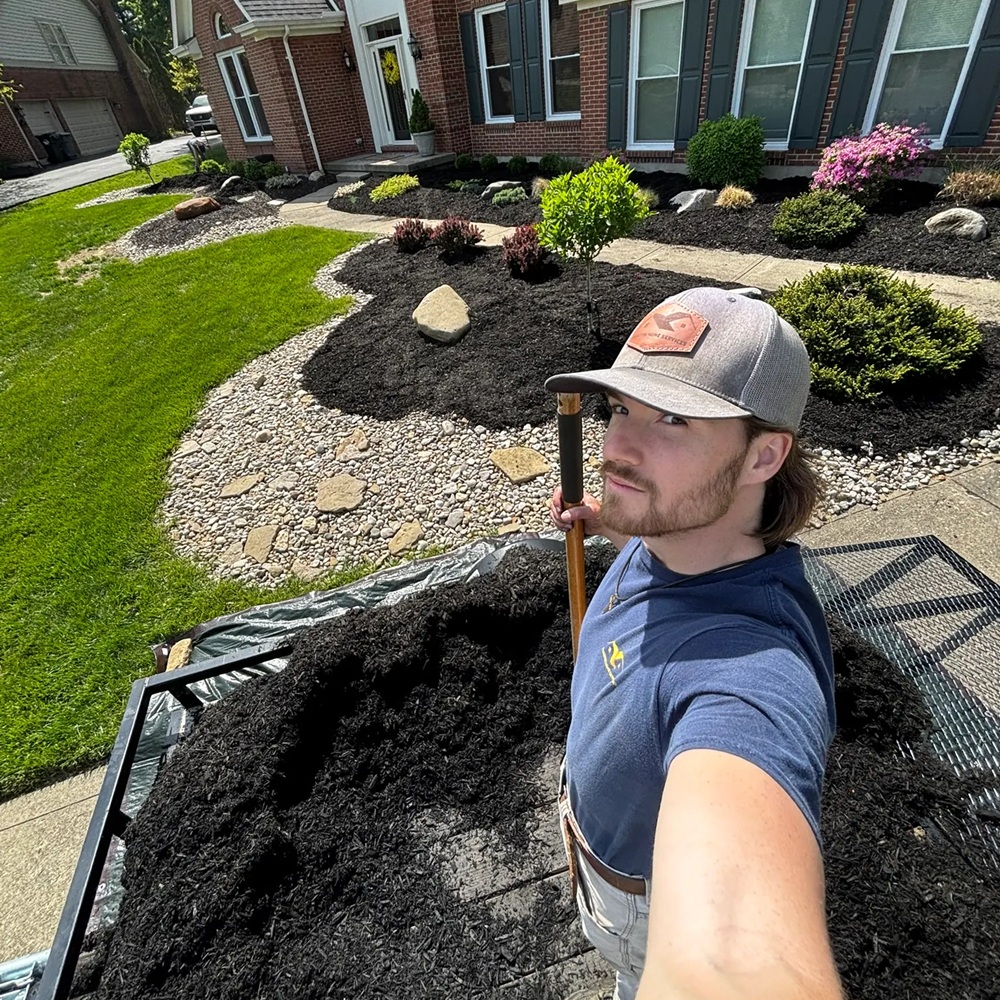Smiling worker taking a selfie in front of new black mulch beds, a stone border and green lawn at a brick home