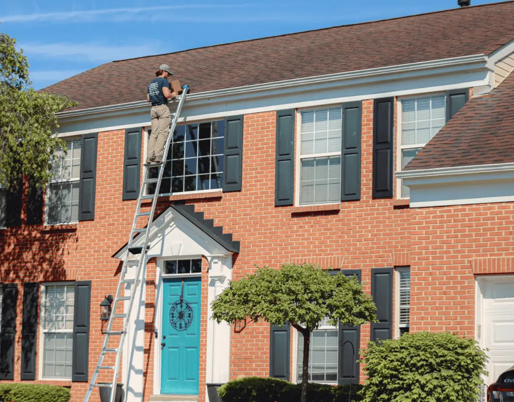 Technician on a tall ladder cleaning the gutters of a two-story red brick home with a teal front door
