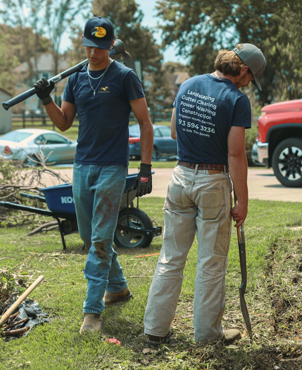 Two landscapers carrying a shovel and pickaxe through a yard with a wheelbarrow and cleared brush