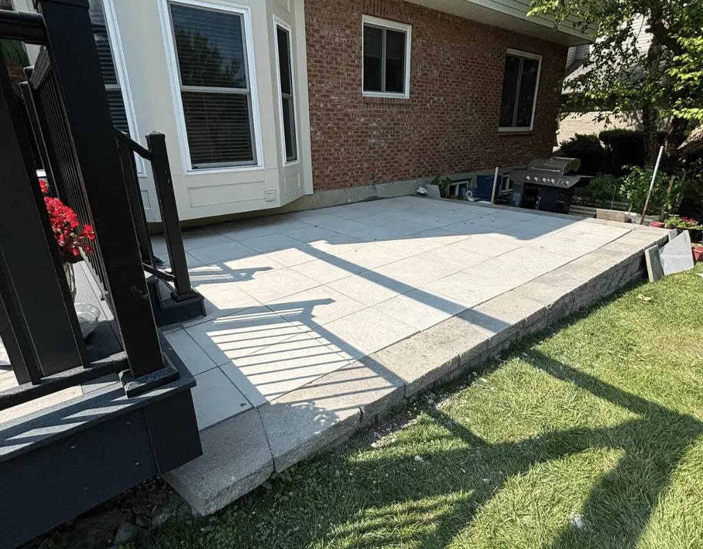 Light gray paver patio extending from the back of a brick home beside a green lawn